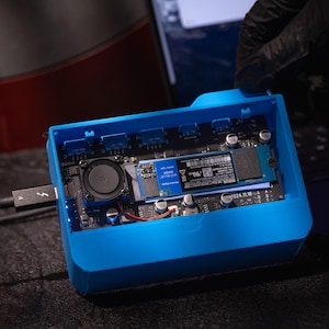 Könnte beinhalten: Ein leuchtend blaues externes Festplattengehäuse mit einer schwarzen internen Leiterplatte, einem kleinen Lüfter und einer blauen Solid-State-Festplatte. Ein schwarzes Kabel ist angeschlossen. Das Gerät ist geöffnet und zeigt seine internen Komponenten.