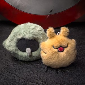 May include: Two fluffy plush toys. One is pale green with a black oval and grey beak. The other is tan with a smiling face, red tongue, and small ears. Both are on a dark surface.