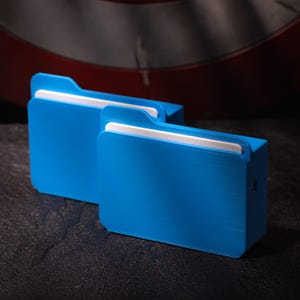 May include: Two bright blue, rectangular storage containers with white accents. The containers have a file folder design, with a tab at the top. The containers are made of a solid material and are placed on a dark surface.