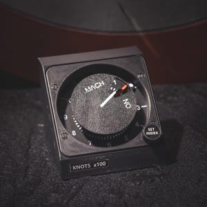 May include: A black aircraft instrument with a round dial displaying "MACH" and "KNOTS x100". The dial has a white needle and numbers, with a small button labelled "SET INDEX". The instrument is likely used for measuring speed.