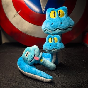 May include: Three plush blue snake toys with yellow eyes and pink tongues. One snake is coiled, another is upright, and the third is in between. The background includes a Captain America shield.