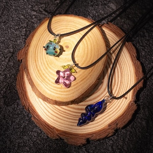 May include: Three glass pendant necklaces with black cords rest on a wooden surface. The pendants are a blue and green apple, a pink flower, and a blue spiral. Each pendant has a silver-colored bail.