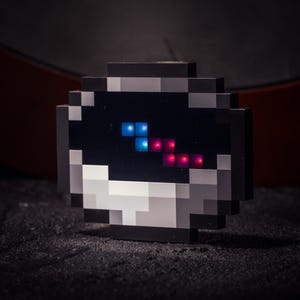 May include: A pixelated, black and gray decorative object with a dark screen displaying blue and red illuminated squares. The object has a retro video game aesthetic, with a square shape and blocky design. The background is dark.