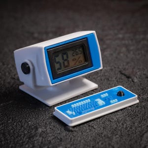 May include: A white and blue miniature computer-like device with a digital display showing numbers and percentages. A separate blue and white rectangular piece with a keyboard-like design is also present. The device has a black button on the side.