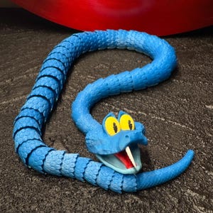 May include: A blue articulated snake toy with a cartoonish design. The snake has yellow eyes, a red mouth, and a white fang. The body is segmented, allowing for flexible movement. The toy is made of plastic and sits on a dark, textured surface.