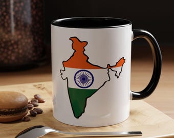 Op de afbeelding: Witte keramische mok met een zwart interieur en handvat. De mok heeft een kleurrijke omtrek van India, met het ontwerp van de Indiase vlag. Een lepel, een macaron en koffiebonen liggen op een houten oppervlak.