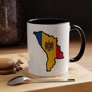 May include: White ceramic mug with a black interior and handle. The mug features a colourful outline of the Moldova map, filled with the country's flag colours. The mug is on a wooden surface with a spoon and a pastry.