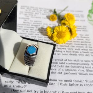 May include: A silver ring with a geometric design, featuring a hexagonal turquoise stone. The ring is displayed in a black jewellery box, with yellow flowers and text in the background.