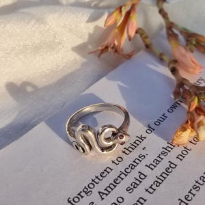 Könnte beinhalten: Ein silberner Schlangenring mit einem roten Edelsteinauge. Der Ring liegt auf einer Seite mit dem Text "forgotten to think of our own Americans." Ein einzigartiger Schmuck.