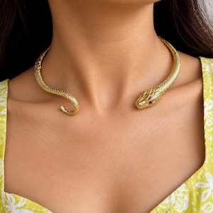 Puede incluir: Un collar tipo gargantilla de metal dorado con forma de serpiente enroscada. El cuerpo texturizado rodea el cuello y presenta una cabeza de serpiente detallada con pequeños ojos rojos incrustados. El diseño es abierto en la parte frontal.