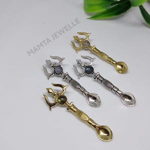 Puede incluir: Cuatro cucharas decorativas con tapas en forma de tridente y pequeños cuencos de cuchara. Dos son doradas y dos plateadas. Cada cuchara tiene una piedra preciosa en el centro del tridente. El texto "MAMTA JEWELLE" es visible.