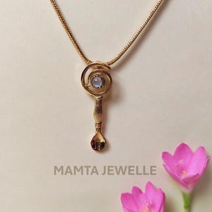 Può includere: Una collana con pendente dorato dal design unico. Il pendente presenta un motivo a spirale con una pietra preziosa azzurra al centro e una forma a cucchiaio nella parte inferiore. La collana è su una catena d'oro. Il testo "MAMTA JEWELLE" è in basso.