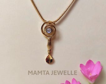 May include: A gold-colored pendant necklace with a unique design. The pendant features a spiral design with a light blue gemstone in the center, and a spoon-like shape at the bottom. The necklace is on a gold chain. The text "MAMTA JEWELLE" is at the bottom.