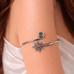 May include: Silver-toned arm cuff featuring a sunburst design with a central amber-colored stone. The cuff has a spiral detail at one end and a textured pattern near the sunburst. This arm cuff is a unique piece of jewelry.