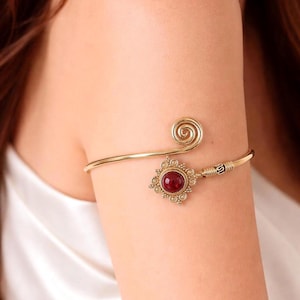 May include: Gold-toned arm cuff featuring a spiral design and a decorative element with a red gemstone. The cuff is worn on the upper arm and has an ornate, vintage aesthetic. The gemstone is a deep red color, set in a detailed, sunburst-like setting.