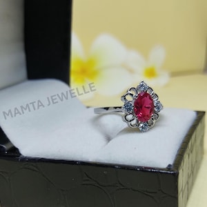 May include: A silver ring with a large oval ruby gemstone at the center, surrounded by smaller blue gemstones. The ring is set in a decorative floral design. The ring is displayed in a black jewelry box with the text "MAMTA JEWELLE".