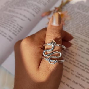 Può includere: Un anello color argento a forma di serpente, con squame dettagliate e un corpo arrotolato. L'anello è indossato su un dito, con la testa del serpente in alto. Un gioiello.