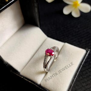 Peut inclure: Une bague en argent ornée d'une pierre précieuse ovale rouge vif, ornée de petites pierres claires le long de la bande. La bague est présentée dans un écrin noir avec un intérieur blanc. Le texte "MAMTA JEWELLE" est visible.