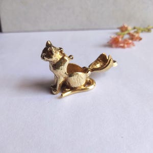 May include: A gold-coloured, cat-shaped trinket box with an open tail. The cat is sitting with its front paws forward. The box is made of a shiny metal and has a small loop at the top for a chain. The tail opens to reveal a small compartment.