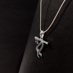 Könnte beinhalten: Eine silberfarbene Anhängerkette mit einem flötenförmigen Anhänger, der ein geschwungenes, bandartiges Design aufweist und an einer feinen Schlangenkette hängt. Das Metallstück hat eine polierte Oberfläche und ist an einer schlichten Kette befestigt.