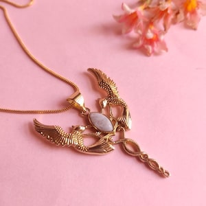 Op de afbeelding: Goudkleurige hangerketting met een gedetailleerd ontwerp van twee vogels met uitgespreide vleugels, die een centrale ovaalvormige, lichtgekleurde steen flankeren. De ketting heeft een fijne ketting en is geplaatst tegen een zachtroze achtergrond.