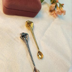 Puede incluir: Dos cucharas pequeñas de metal con un diseño de cabeza de cobra en la parte superior del mango. Una cuchara tiene un acabado en tono plateado pulido, mientras que la otra tiene un acabado en tono dorado pulido. Ambos mangos tienen escamas.