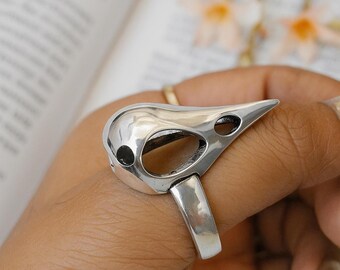 Puede incluir: Un anillo plateado con forma de cráneo de pájaro. El anillo presenta un pico puntiagudo y dos cuencas oculares. El anillo se lleva en un dedo, con un libro y flores en el fondo. El anillo es de metal brillante.