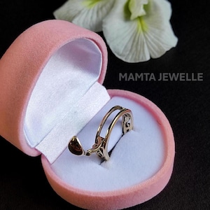May include: A gold ring with an open design, presented in a pink velvet box. The ring has detailed filigree patterns and a small, teardrop-shaped charm. The text "MAMTA JEWELLE" is visible in the background.