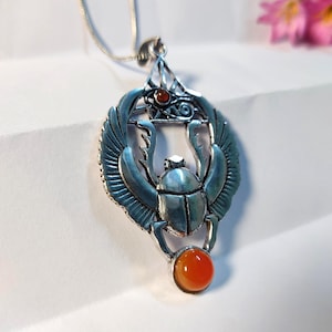 May include: A silver-toned metal pendant necklace featuring a winged scarab beetle design. The pendant includes a small, round, orange-red gemstone at the bottom and a similar smaller stone set within an Eye of Horus symbol at the top.