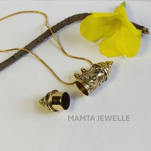 Puede incluir: Un collar con colgante dorado con un dije cilíndrico ornamental y abrible. El dije presenta patrones grabados y una parte superior cónica. El collar se muestra con una flor amarilla y una pequeña rama. El texto "MAMTA JEWELLE" es visible.