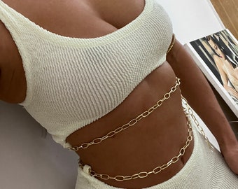 May include: A white ribbed crop top and matching skirt with a gold chain belt. The chain belt is layered and wraps around the waist.
