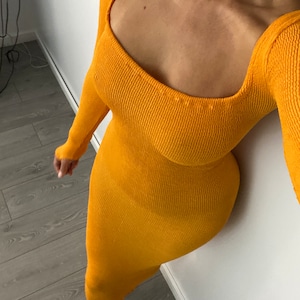 Può includere: Un abito lungo a maniche lunghe di colore senape con scollo quadrato. L'abito è realizzato in tessuto a costine.