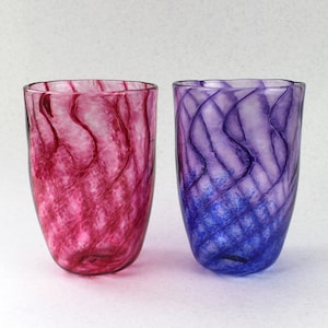 May include: Two glass tumblers with swirling patterns. The tumbler on the left is pink with red swirls, and the tumbler on the right is purple with blue swirls.