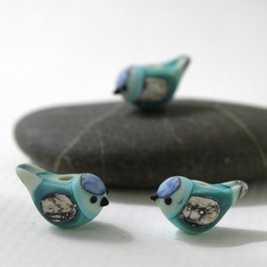 May include: Three glass bird figurines in shades of teal, blue, and white. The birds have black beaks and are perched on a dark gray stone. The figurines have a unique, handcrafted appearance, suggesting they are decorative items.