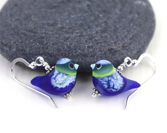 Könnte beinhalten: Ein Paar blaue Vogelohrringe mit silbernen Haken und weißen und grünen Details. Die Vögel sind aus Glas und haben eine strukturierte Oberfläche.