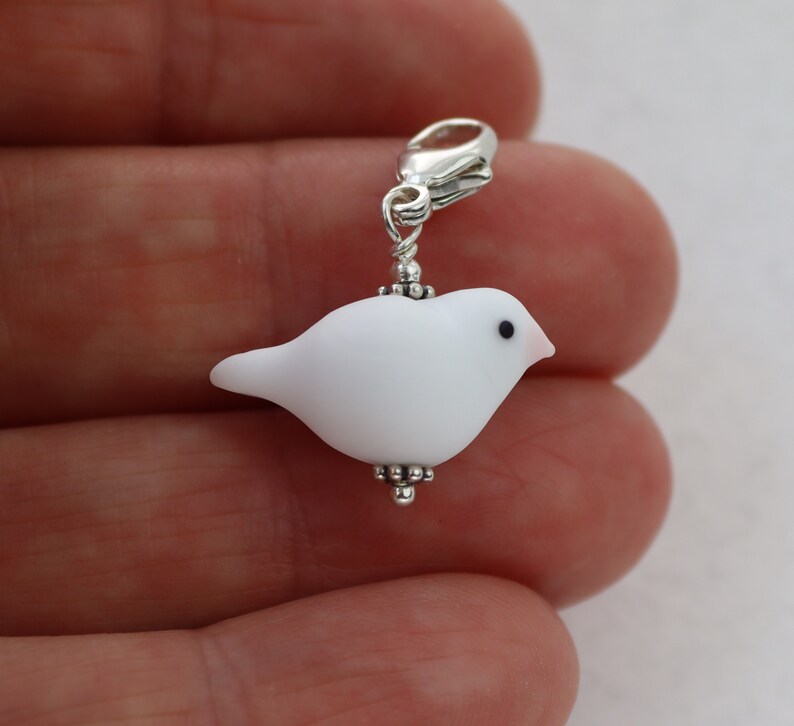 May include: White glass bird charm with a silver clasp. The bird has a black eye and a pink beak.