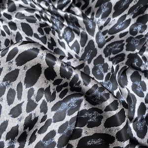 Może przedstawiać: Zbliżenie tkaniny z wzorem w cętki. Tkanina ma satynowy połysk i czarne i szare plamy na jasnoszarym tle. Tkanina jest udrapowana, ukazując fałdy i cienie.