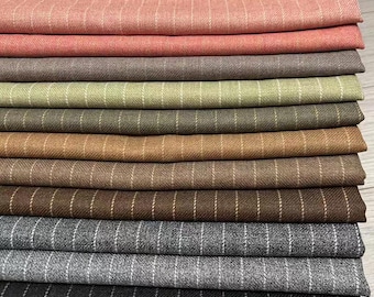 May include: A selection of pinstripe fabrics in a range of colours, from pale pink to black. The stack includes shades of grey, brown, and green. The fabrics are neatly folded, displaying the different textures and hues.