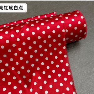 Peut inclure: Un rouleau de tissu rouge &agrave; pois blancs. Le tissu est pli&eacute;, r&eacute;v&eacute;lant le motif et le bord roul&eacute;. Le texte "亮红底白点" est visible dans le coin sup&eacute;rieur gauche.