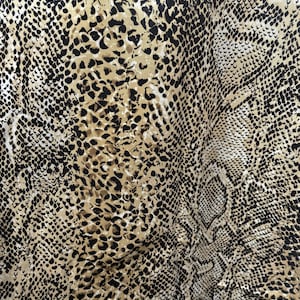 Op de afbeelding: Close-up van een stof met een slangenprint. Het ontwerp combineert zwart, bruin en crèmekleurige tinten, die de uitstraling van reptielenhuid nabootsen. De stof lijkt zacht en soepel, met zichtbare plooien en texturen.