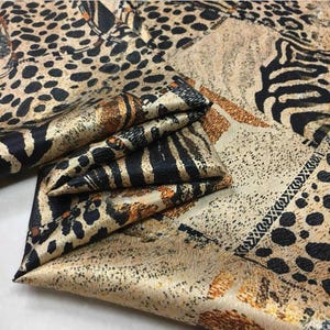 Könnte beinhalten: Ein gefalteter Stoff mit einem Patchwork-Design mit Tiermotiven. Der Stoff zeigt Muster von Leopardenflecken, Zebrastreifen und einem gesprenkelten Hintergrund in Braun-, Schwarz- und Beigetönen. Der Stoff hat einen seidigen Glanz.