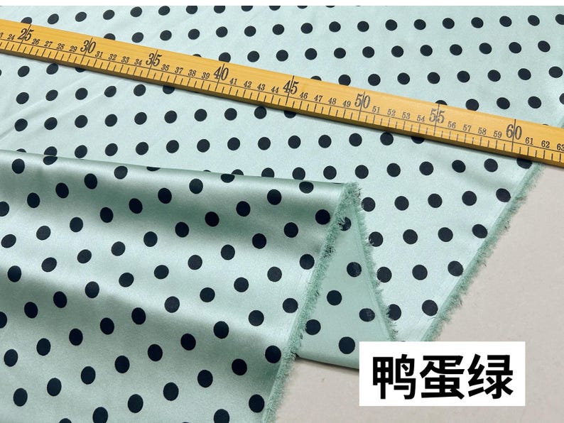 Peut inclure: Un tissu vert clair avec un motif r&eacute;p&eacute;t&eacute; de pois noirs. Une r&egrave;gle en bois avec des mesures en centim&egrave;tres est pos&eacute;e sur le tissu. Le tissu a une texture lisse, semblable &agrave; du satin. Le texte "鸭蛋绿" est dans un encadr&eacute; blanc en bas &agrave; droite.