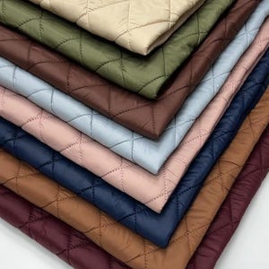 Könnte beinhalten: Ein Stapel gesteppter Stoffmuster in verschiedenen Farben, darunter Beige, Olivgrün, Braun, Hellblau, Rosa, Marineblau und Burgunderrot. Jedes Stück hat ein durchgenähtes Rautenmuster.