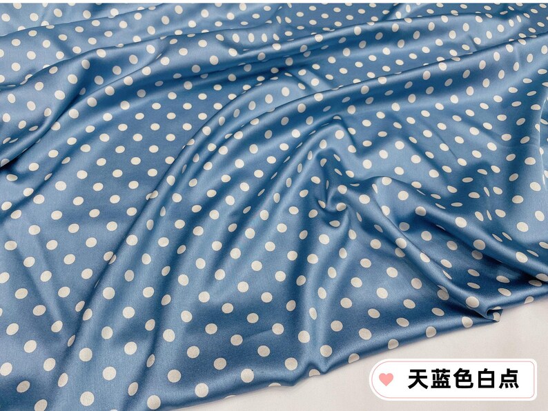 Peut inclure: Gros plan sur un tissu bleu avec des pois blancs. Le tissu est drap&eacute;, cr&eacute;ant des plis et des ombres douces. Le tissu a un aspect brillant, semblable &agrave; du satin. Le texte sur l'image dit "Polka Dots Bleu Ciel".