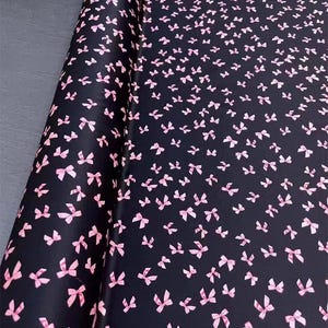 Puede incluir: Tela negra con un patrón repetido de pequeños lazos rosas. Los lazos están dispersos por la superficie, creando un diseño delicado y femenino. La tela está enrollada, mostrando la textura y el patrón.