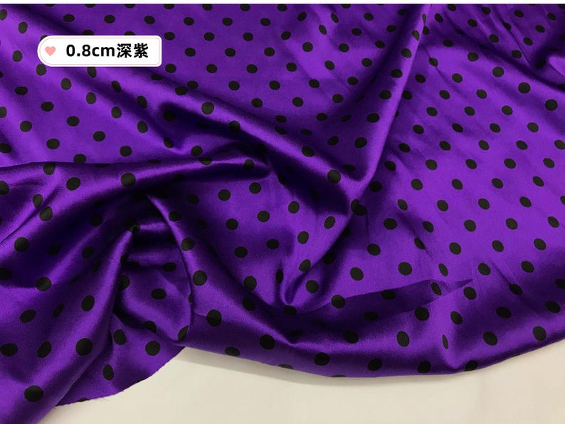Peut inclure: Un tissu violet vif avec un aspect brillant, orn&eacute; de pois noirs espac&eacute;s r&eacute;guli&egrave;rement. Le tissu est pli&eacute;, cr&eacute;ant des ombres et des reflets. Le texte sur l'image indique "0,8 cm".