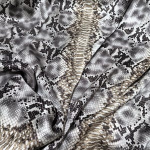 Może przedstawiać: Zbliżenie tkaniny ze wzorem skóry węża. Tkanina ma powtarzający się wzór szarych, czarnych i białych łusek, z centralnym pasem w kolorze brązowym i brązowym. Tkanina jest udrapowana, ukazując fałdy i fakturę.