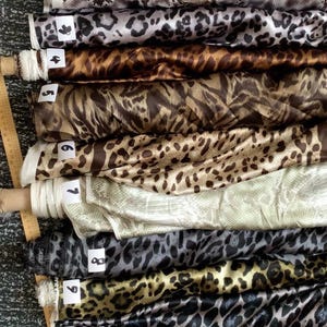 Może przedstawiać: Kolekcja rolek materiału z różnymi wzorami zwierzęcymi, w tym wzory węża, lamparta i tygrysa. Tkaniny są w odcieniach brązu, czerni, szarości i złota. Każda rolka jest oznaczona numerem.