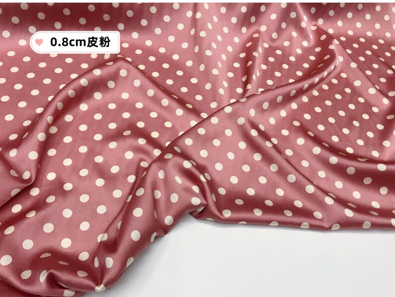 Peut inclure: Gros plan sur un tissu satin&eacute; rose avec un motif &agrave; pois. Le tissu est de couleur rose poudr&eacute; avec des pois blancs. Le texte "0,8 cm 皮粉" est dans le coin sup&eacute;rieur gauche. Le tissu est drap&eacute; et pli&eacute;.