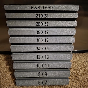Op de afbeelding: Een stapel grijze gereedschappen met de tekst "E&S Tools" en afmetingen in centimeters. De maten variëren van 15,24 cm x 17,78 cm tot 53,34 cm x 58,42 cm. De gereedschappen zijn op elkaar gestapeld.